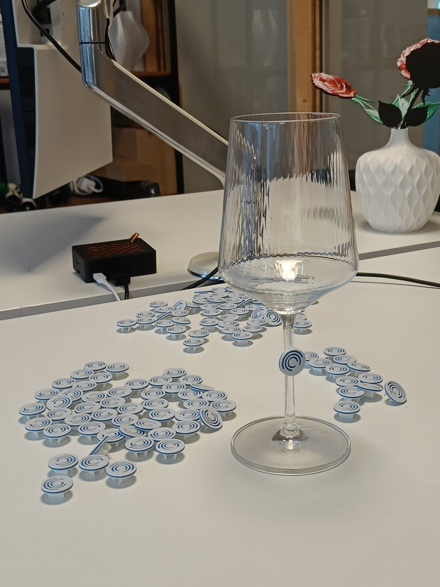 Tientallen ronde witte en blauwe logoclips op een tafel, met één clip aan de steel van een wijnglas