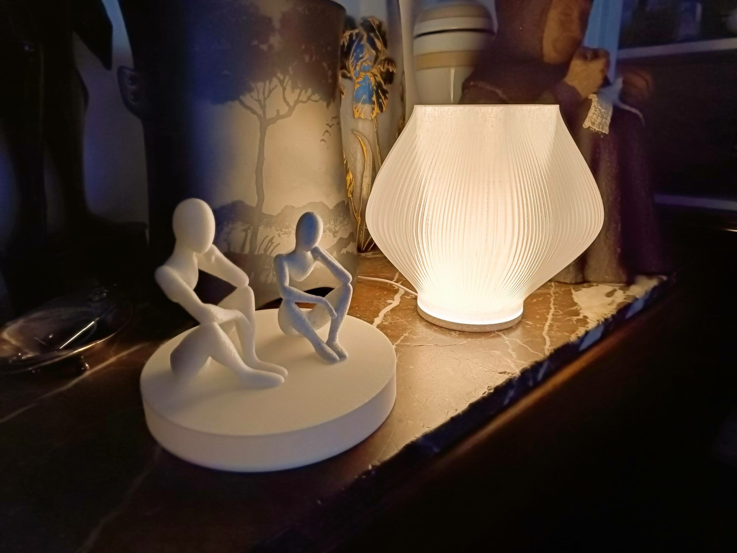 Een gloeiende geribbelde witte lamp en een sculptuur van twee zittende figuren op een ronde voet, allebei 3D-geprint, op een marmeren schouw